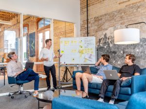 Four men in a conference room. One man is standing presenting his ideas and goals using a whiteboard and pointing with a pen. The other three men are sitting and visibly listening to him present.