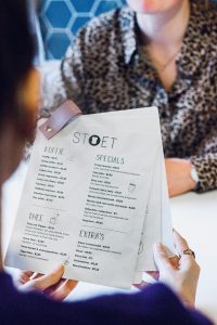 A restaurant menu genre. It is white with modern sans-serif fonts. The hands of one woman holding the papers are visible, while another woman’s arms are across the table.